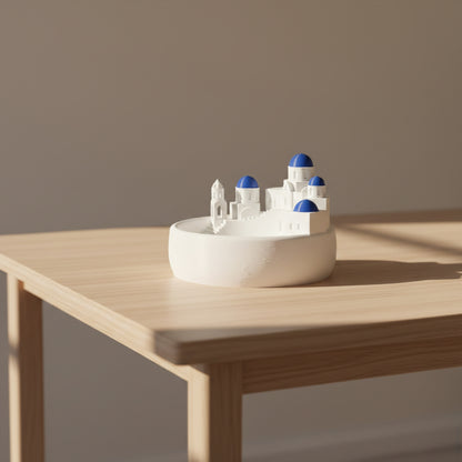 A white 3D-printed miniature model of a Mediterranean-style cityscape with white buildings and blue domes, displayed on a white circular base against a light gray background.